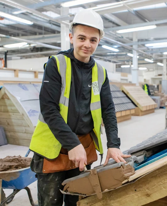 A roofing student smiling while applying mortar to a roof edge, with various roof structures visible in the background of the workshop. A roofing student smiling while applying mortar to a roof edge, with various roof structures visible in the background of the workshop.